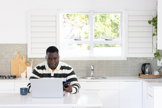 African American man holding smartphone beside open laptop and blue mug at kitchen counter