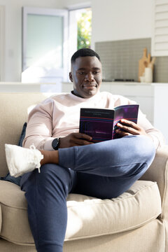 African American man sitting on beige sofa and holding purple booklet while reading at home
