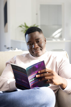 African American man sitting on light-colored couch holding open book while reading in bright room