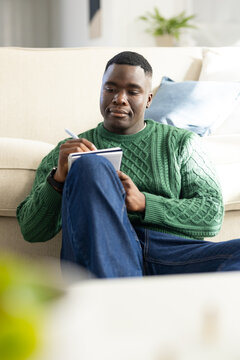 African American man in green sweater sitting on sofa holding notebook and writing with pen