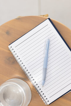 Open spiral notebook lying diagonally on wooden table beside blue pen and glass cup, copy space