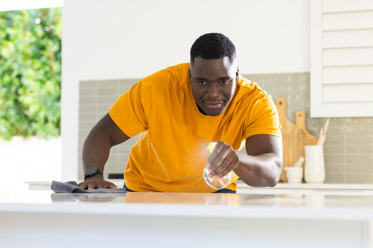 African American man in t-shirt leaning while wiping counter with cloth and spray bottle