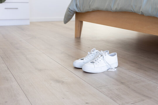 White lace-up canvas sneakers resting on light wood floor beneath bed with gray bedding, copy space