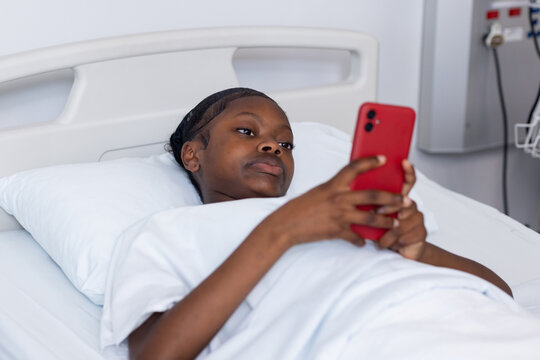 African American teenage girl lying in hospital bed holding red smartphone in both hands