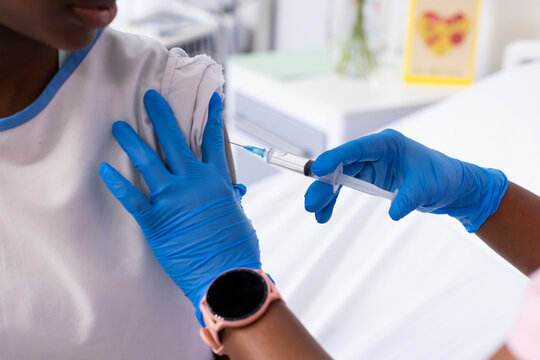 African American woman in blue gloves vaccinating teenage girl upper arm with syringe in clinic