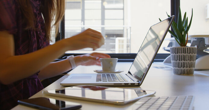 Mid section of young businesswoman drinking coffee while using mobile phone at desk in office