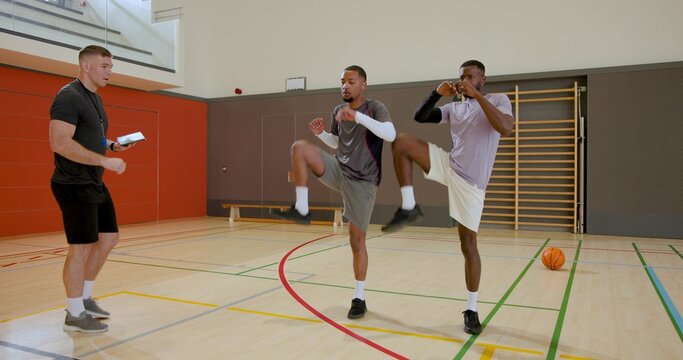 Diverse coach and athletes talking and doing knee drills in sports hall, with tablet and basketball