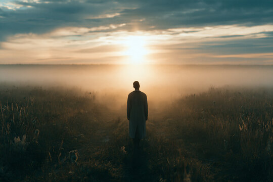 A lone figure in a long coat stands in a vast foggy field at sunset facing the radiant glow on the horizon creating a poignant scene of contemplation and solitude in the golden light
