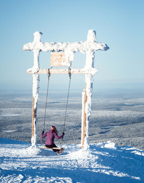 Young woman swinging on a giant snow-covered wooden swing on top of a mountain, scenic winter landscape and winter holiday concept