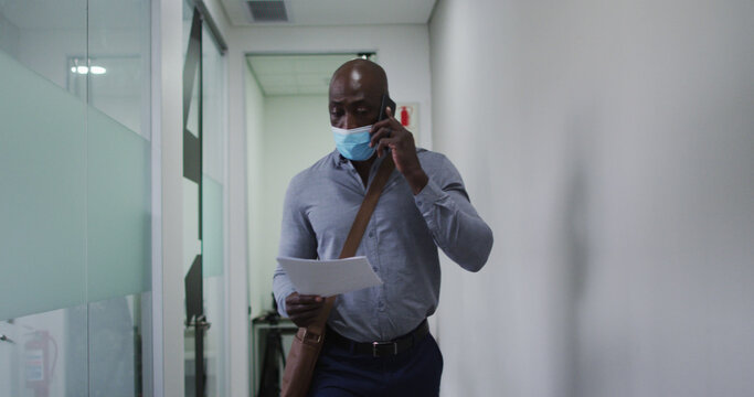 African american businessman in face mask holding document talking on smartphone in office corridor