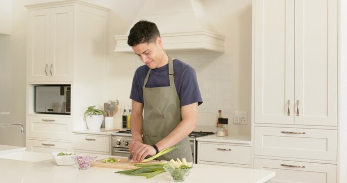 Slicing man wearing olive apron chopping scallions on cutting board in modern kitchen with bowls
