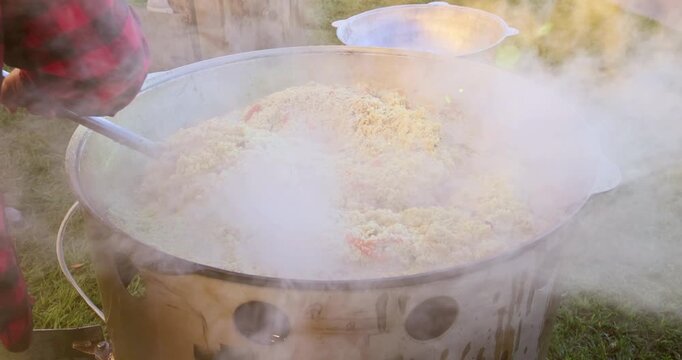 Steam rising from large pot of rice mixed with vegetables being stirred outdoors