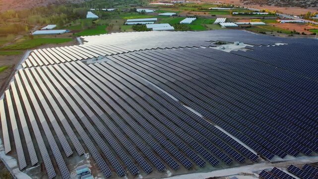 Aerial view of a vast solar power plant with long rows of photovoltaic panels reflecting warm sunlight. Rural fields and farm buildings surround the large renewable energy facility in the countryside.