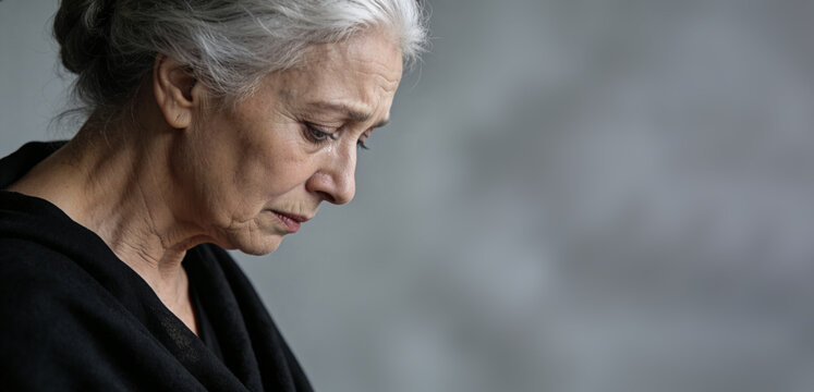 Senior woman with gray hair crying, tear rolling down cheek in close-up profile against gray backdrop. Portrait of elderly woman in grief and sadness. Loss, mourning and sorrow concept.