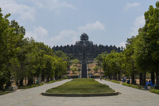 MAGELANG, INDONESIA - OCTOBER 21, 2026: Visitors rest along the tree-lined approach to Borobudur Temple, with its stone terraces and scaffolded top stupa rising above the park in Central Java.
