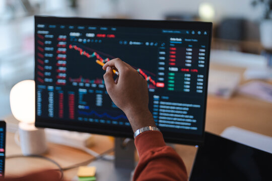 A financial analyst points at a computer screen displaying stock market charts and financial data. The professional is tracking market trends in an office.