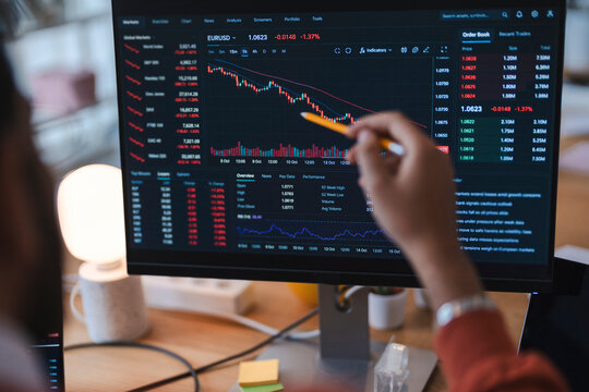 A professional financial analyst points a yellow pencil at a computer monitor displaying a declining stock market graph. The worker reviews falling trading data in the office.