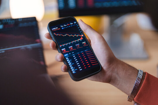 A young investor holds a smartphone to check financial markets. The mobile screen displays red stock market charts and falling graphs during a trading session indoors.