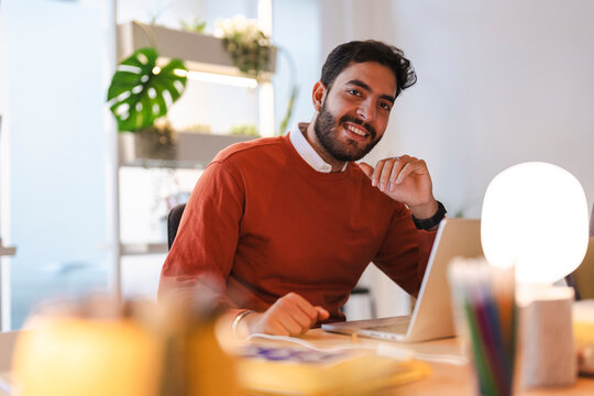 A young bearded man wearing an orange sweater is sitting at a wooden desk with a laptop in a brightly lit modern office, smiling warmly while looking at the camera.