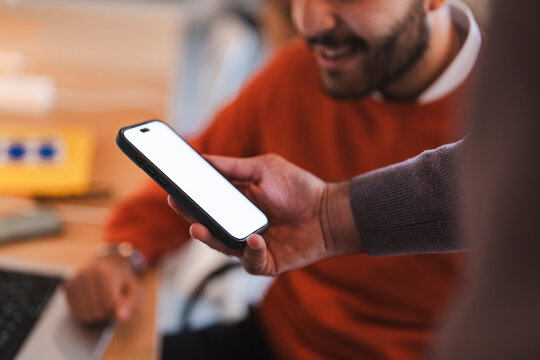 Two colleagues collaborating closely, as a smiling man in a red sweater holds up a smartphone with a blank white screen to share content during an office meeting.
