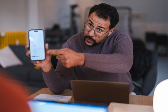 A man with glasses and a beard sits at a desk, holding up a smartphone and pointing at the screen while explaining something to a colleague in a modern office.