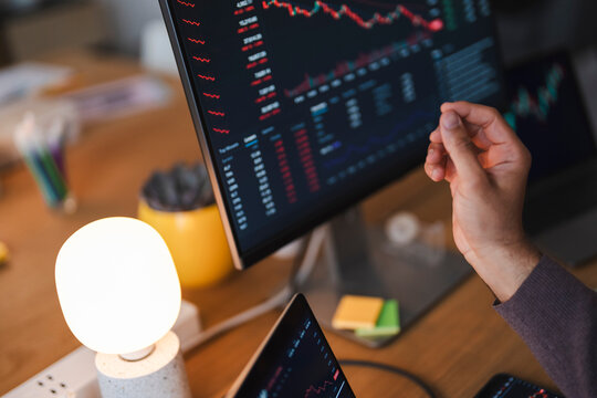 A professional trader analyzes stock market charts and falling financial data trends on multiple computer monitors in a bright office. He points at the screen.