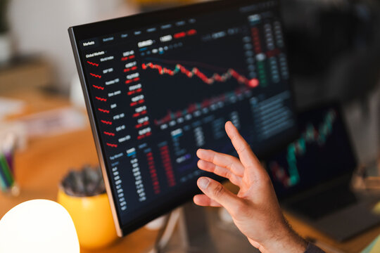A financial trader points at a computer monitor displaying stock market charts and red data graphs in an office. They analyze market trends during a trading session.