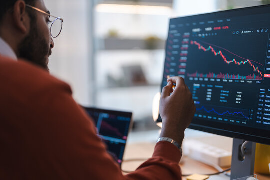 A focused male trader wearing glasses sits at his desk, carefully analyzing financial stock market charts and trading data on multiple computer screens in a modern office.