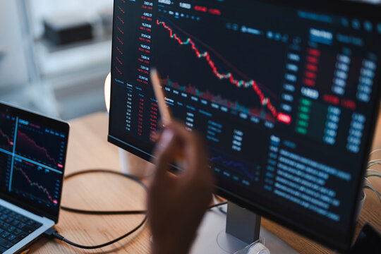 A financial analyst points at a computer monitor displaying stock market charts. The trader reviews negative financial trends in the global market from an office.