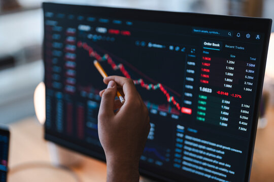 A financial analyst holds a pencil and points at stock market charts and trading data displayed on a computer screen, closely reviewing financial trends and market movements.