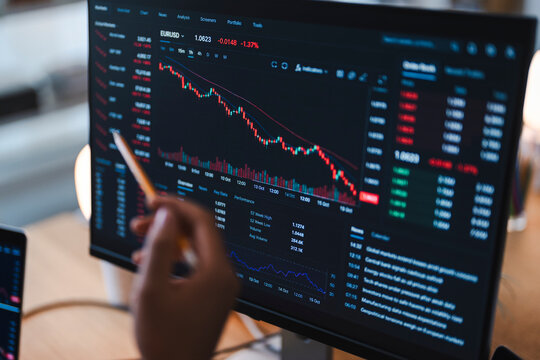 A financial analyst points at a computer monitor displaying falling stock market charts and negative trading data. The graphs show a significant downward trend in the market.