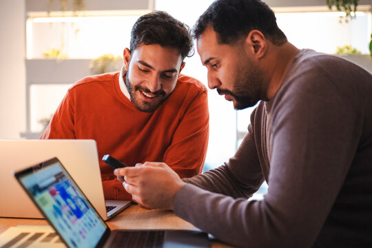 Two male business colleagues collaborate in a bright modern office. They look at a smartphone together while sitting at a wooden desk with open laptops.