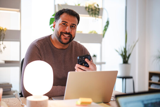 A smiling man with a beard sits at an office desk holding a smartphone. He looks directly at the camera with a friendly expression next to his laptop and lamp.