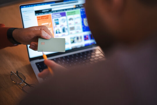 A person hands a credit card to someone sitting at a wooden desk with an open laptop computer. They are making an online purchase together in a modern office room.
