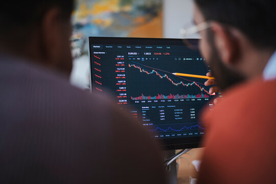 Two male colleagues analyze financial stock market data on a computer screen. One man points a pencil at the declining chart trends while discussing business strategy together.