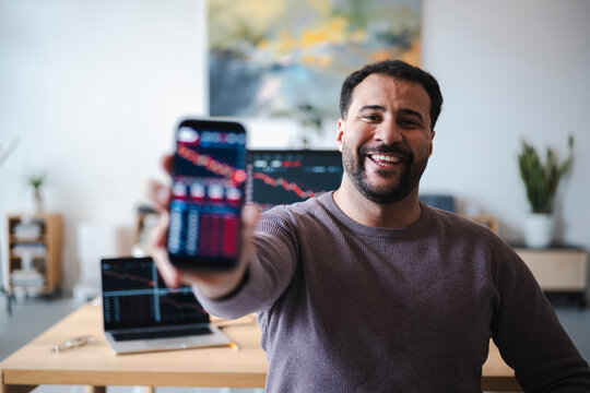 A smiling man in a sweater sits at a desk and holds up a smartphone showing financial market charts. Computer screens with stock trading data are visible in the background.