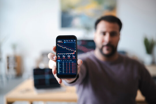 A man holds a smartphone displaying a stock market trading app with declining charts. He shows the financial data and red graphs to the camera in a bright home office.