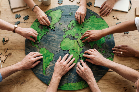 Diverse hands assembling round wooden world map puzzle on rustic table. Top view of group joining green continents together. Global teamwork, unity and collaboration concept.