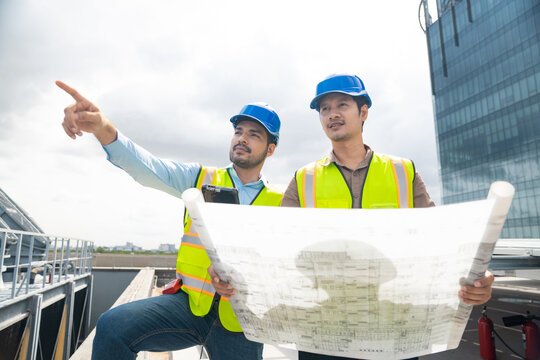 Two engineers with digital tablet and blueprint pointing at building construction site. Professional HVAC engineers using digital tablet and pointing at installation site rooftop facility inspection.