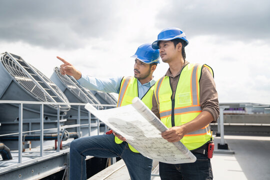 Two engineers with digital tablet and blueprint pointing at building construction site. Professional HVAC engineers using digital tablet and pointing at installation site rooftop facility inspection.