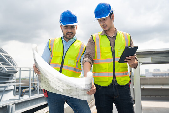 Two engineers holding and examining large building blueprint on rooftop. Construction supervisor and HVAC technician analyzing architecture paper blueprint together on industrial building site.