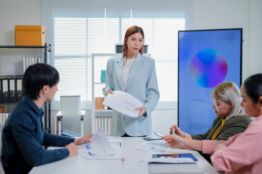 Asian businesswoman leaning over table and pointing at business proposal while guiding team through quarterly report in modern office with AI assistant screen