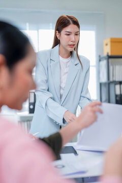 Asian businesswoman leaning over table and pointing at business proposal while guiding team through quarterly report in modern office with AI assistant screen