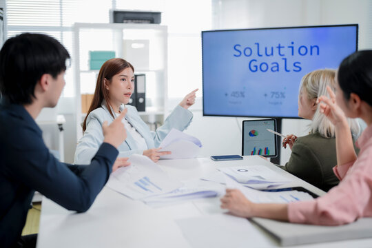 Asian businesswoman leading interactive team meeting and pointing at solution goals on screen while colleagues raise hands and share opinions in office