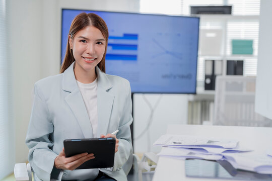 Focused young Asian businesswoman in light blazer reading report on digital tablet in modern office meeting room with presentation screen and copy space