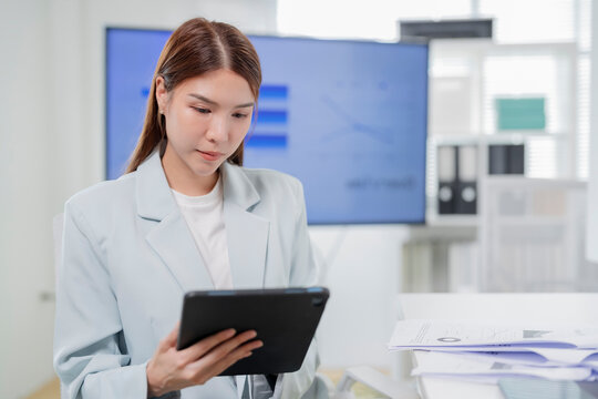 Focused young Asian businesswoman in light blazer reading report on digital tablet in modern office meeting room with presentation screen and copy space