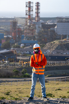 Industrial worker in respirator and safety gear standing before a polluting factory