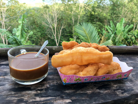 Traditional Thai breakfast style featuring deep-fried dough stick (Pa Thong Ko) and a cup of hot coffee, placed on a wooden table with a scenic green forest background, perfect for travel and local