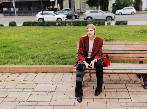 Young Woman Sitting on Bench in City Park With a Leg Brace