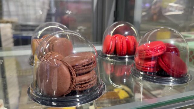 French macaron cookies inside transparent plastic dome packages stand on a bakery shop display shelf. Prepackaged chocolate and strawberry sweet macarons confectionery retail layout close up.
